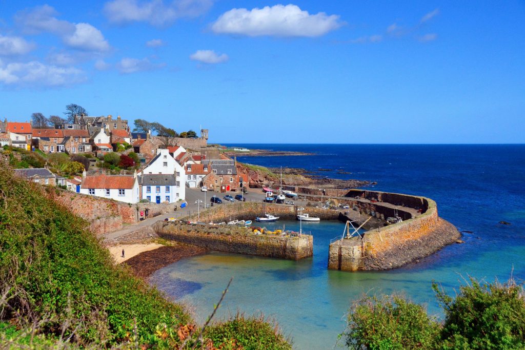 Panoramic view of Crail Harbour, showcasing historic stone buildings, fishing boats docked along the serene water, and the rugged coastline. A perfect destination for those exploring Scotland's beautiful coastal villages.