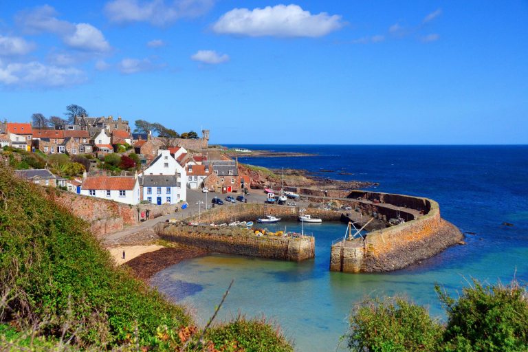 Panoramic view of Crail Harbour, showcasing historic stone buildings, fishing boats docked along the serene water, and the rugged coastline. A perfect destination for those exploring Scotland's beautiful coastal villages.
